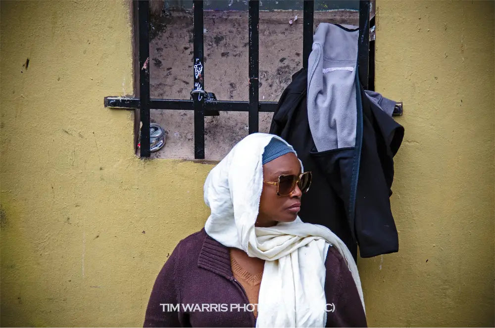 Woman wearing white headscarf and sunglasses in dark purple sweater standing against yellow wall with black metal window and hanging clothes