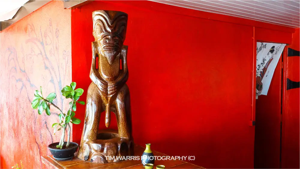 Carved wooden tiki statue with detailed face against bright red wall with small potted green plant and ceramic vessel on shelf