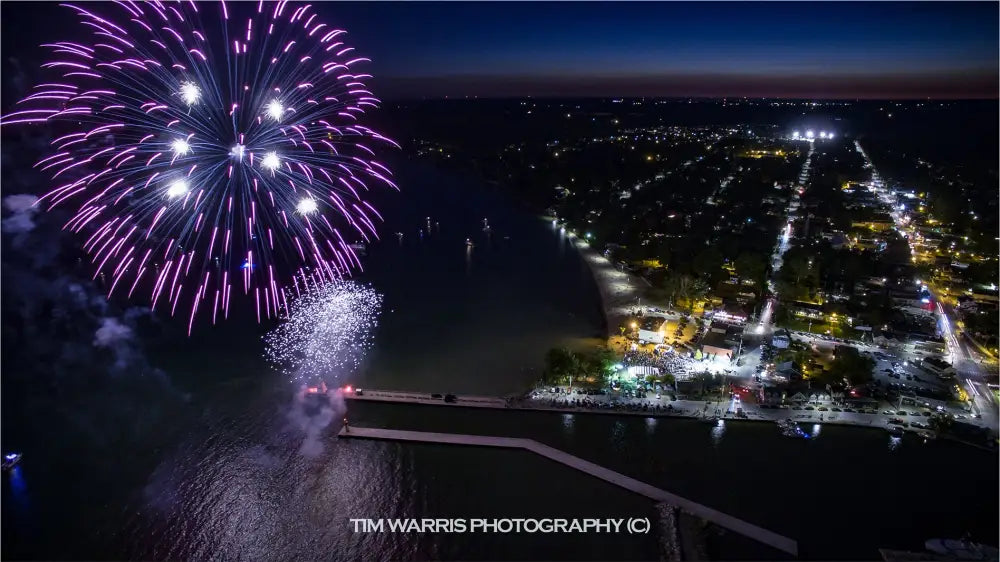 Aerial view of purple and white fireworks bursting over illuminated waterfront town at night with street lights glowing across residential neighborhood under deep blue twilight sky