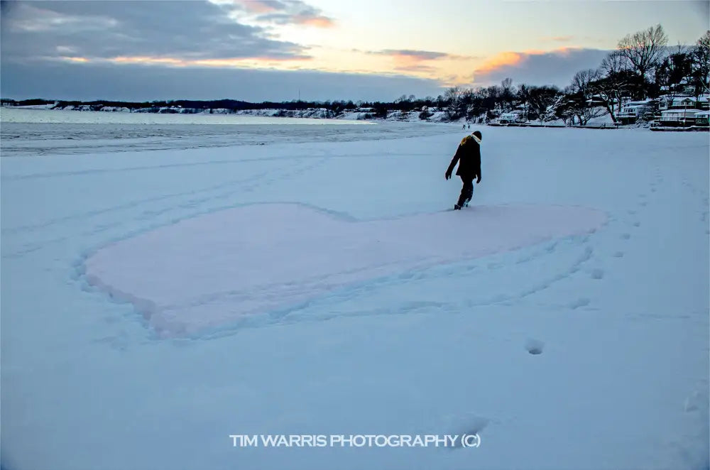 Solitary person in dark winter clothing walking across vast frozen lake with footprints trailing behind under blue winter twilight sky with bare trees and shoreline visible