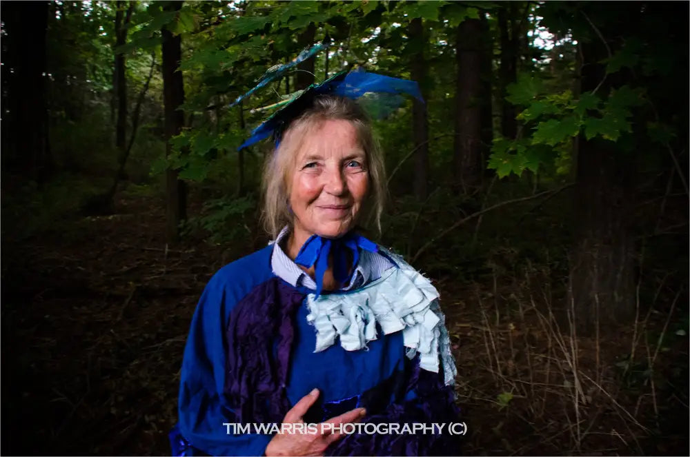 Woman in colonial period costume wearing blue headscarf and blue cape with white and black decorative pattern standing in wooded area
