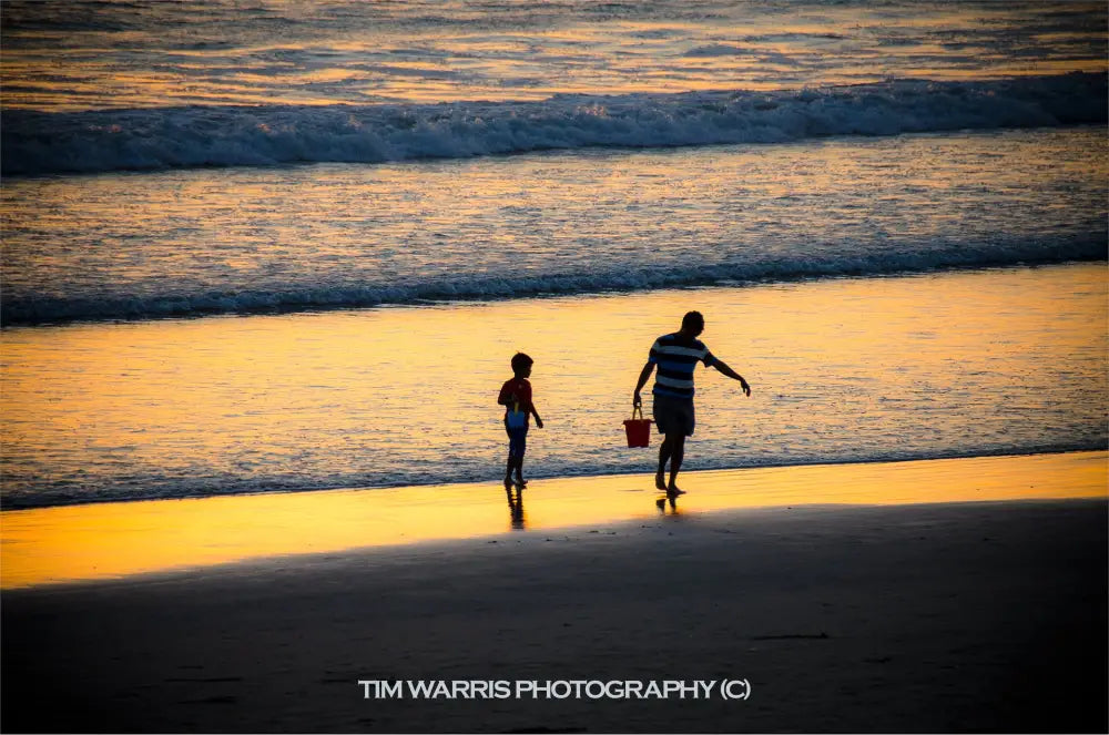 Silhouettes of adult and child walking on wet beach at sunset with golden reflections on sand and ocean waves in warm evening light